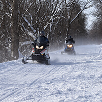 Find the snow Two snowmobile riders on a snowy trail in the woods.